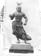 右手を握りしめて左手を広げている金剛力士像の全体像を正面から離れて撮影したモノクロの写真