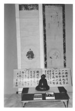 壁に2枚の掛軸が掛かりその前に文字の書かれた木の板が立てかけてありその前に小さな仏像と仏像の前に小さい木箱が置かれている様子を正面から撮影した白黒の写真
