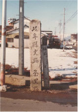 雪が一面に広がる広場の一角に文字が書かれた縦長な石碑が設置されその横に2本石の台に鉄の棒がたっている写真