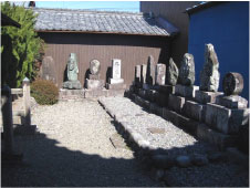 建物に囲まれた砂利のの地面の広場に四角い石が3段積まれた句碑がL字に並べられている写真