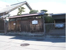 瓦屋根の家とガレージに囲まれた土地に屋根のついた古い門と白い案内板が立っている五竹坊居宅跡の写真