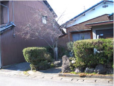 茶色の外壁の一般家屋を左に敷地の外側から梔庵居宅跡を生垣の入口を正面から写した写真