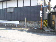 黒い壁の建物の横にあるオレンジのポールのカーブミラーのそばに柱本高札場跡の標柱が立っている写真