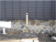 黒い壁の建物の近くにある白いガードレールの近くに柱本高札場跡の標柱が立っている写真