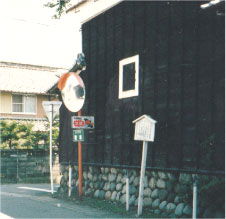 黒い壁の建物のすぐ脇にあるオレンジのカーブミラーに並んで案内板が立っている写真