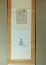 座っている女性の絵と和歌が描かれた茶色の掛け軸を撮影した写真