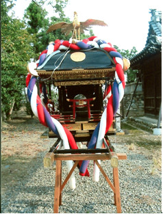 神社の境内にある赤青白のしめ縄で装飾された黒で塗られ屋根に鳳凰の像が設置された神輿の写真