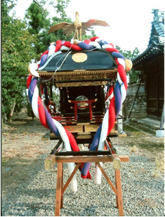 屋外で黒で塗られ赤青白のしめ縄で装飾された屋根に鳳凰像がある神輿を正面から撮影した写真