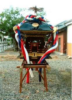 屋外で黒で塗られ赤青白のしめ縄で装飾された屋根に鳳凰像がある神輿を背面から撮影した写真