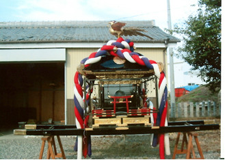 屋外で黒で塗られ赤青白のしめ縄で装飾された屋根に鳳凰像がある神輿を側面から撮影した写真